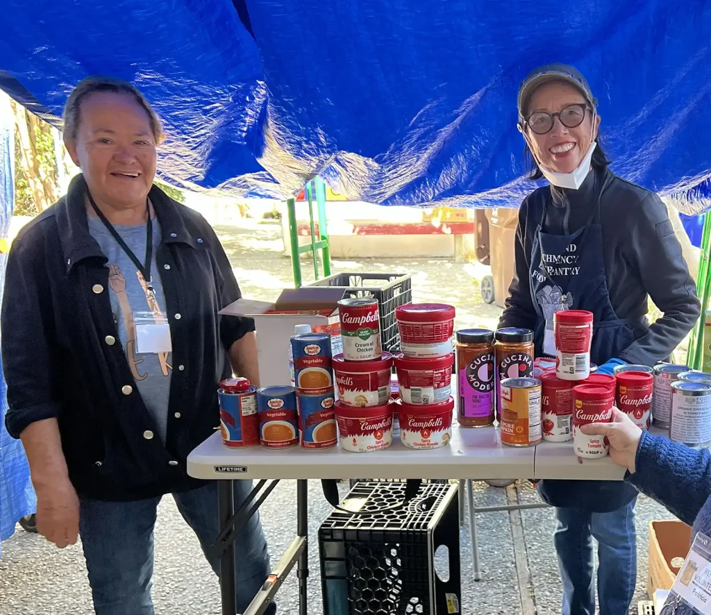 Two volunteer beside table of mostly canned food